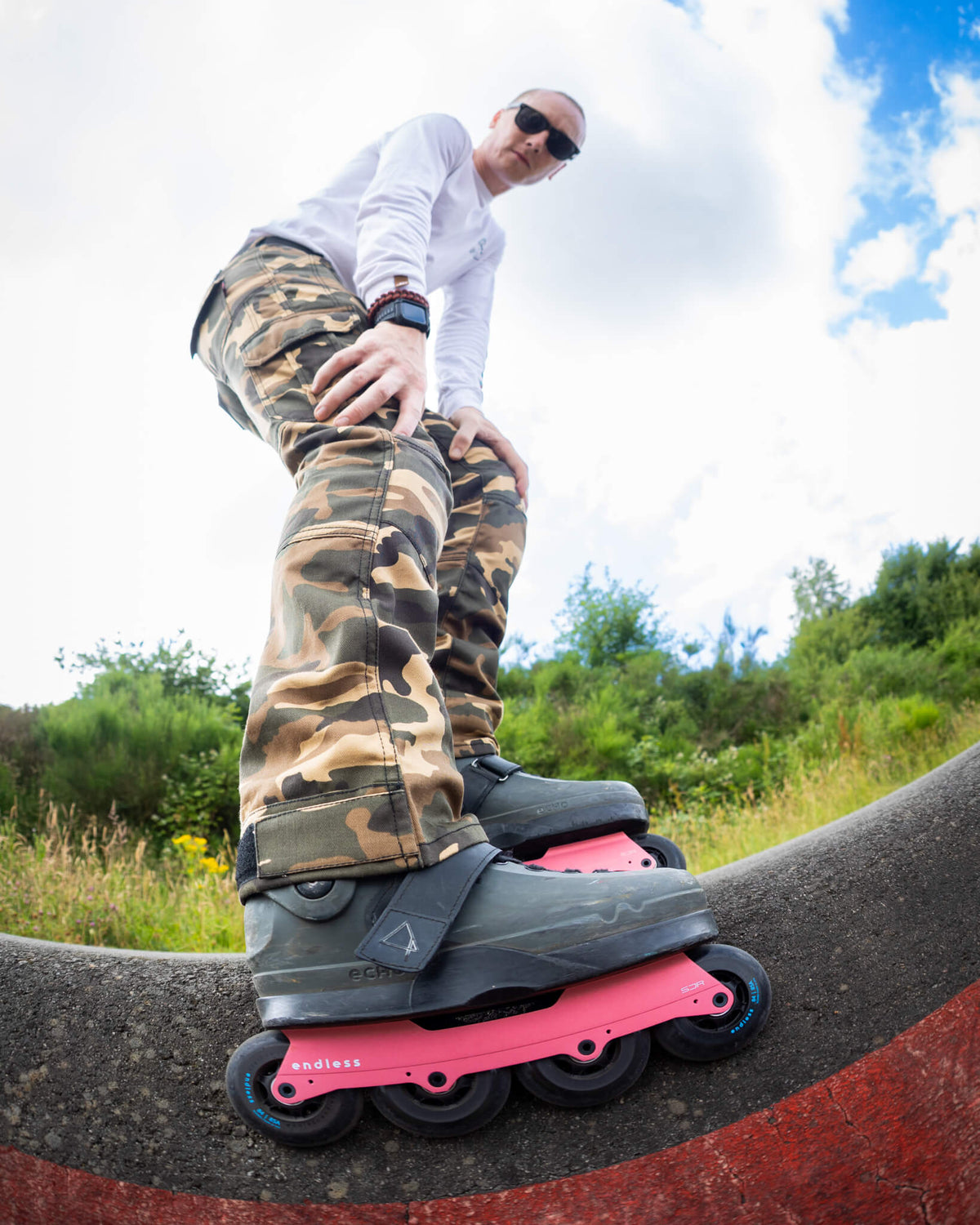 A man wearing sunglasses, a white long-sleeve shirt, and camouflage pants is balancing on a Light Red Endless SJR inline skate outdoors on a curved concrete ramp with grass and trees in the background.