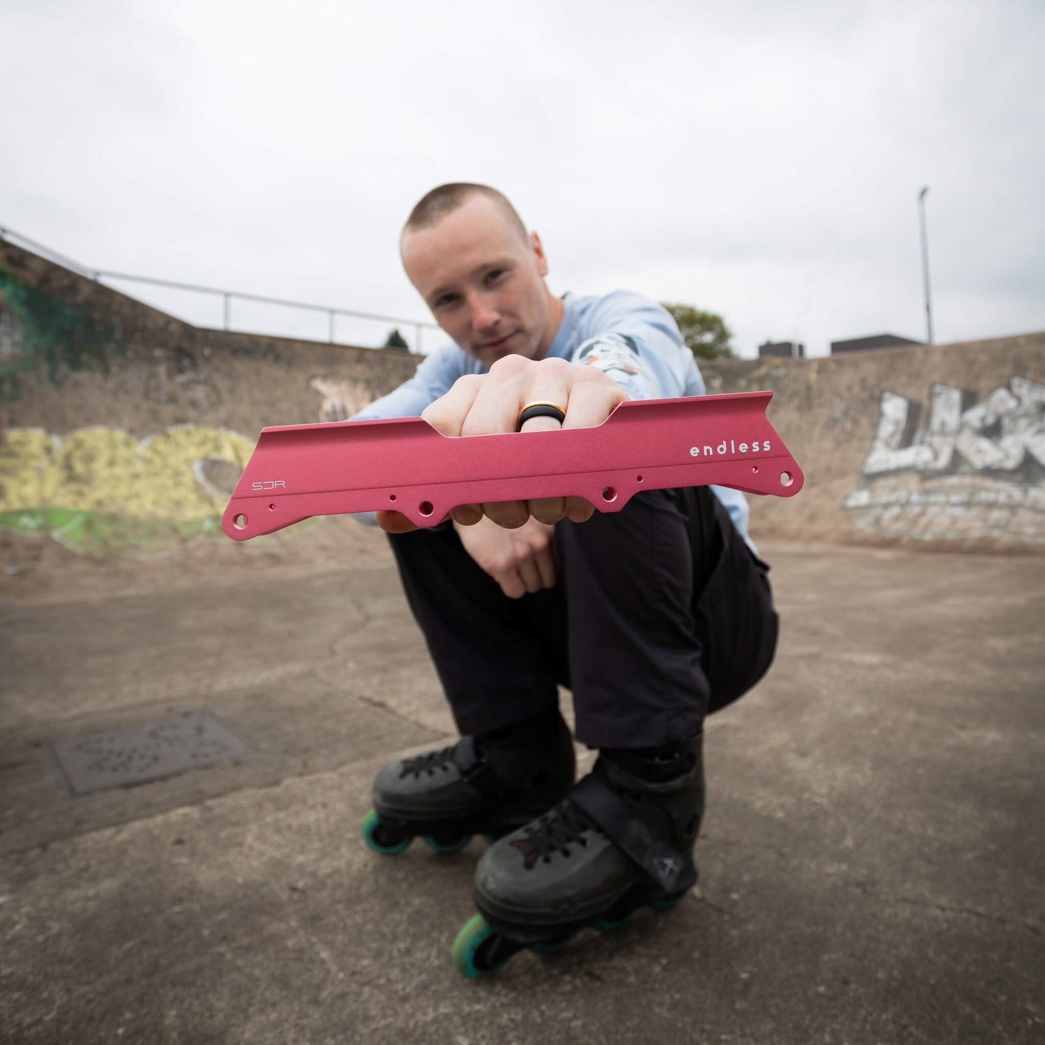 A man in a skatepark crouches down holding a Light Red Endless SJR inline skate frame in front of his face. The frame has the brand name 'endless' printed in white on the side.