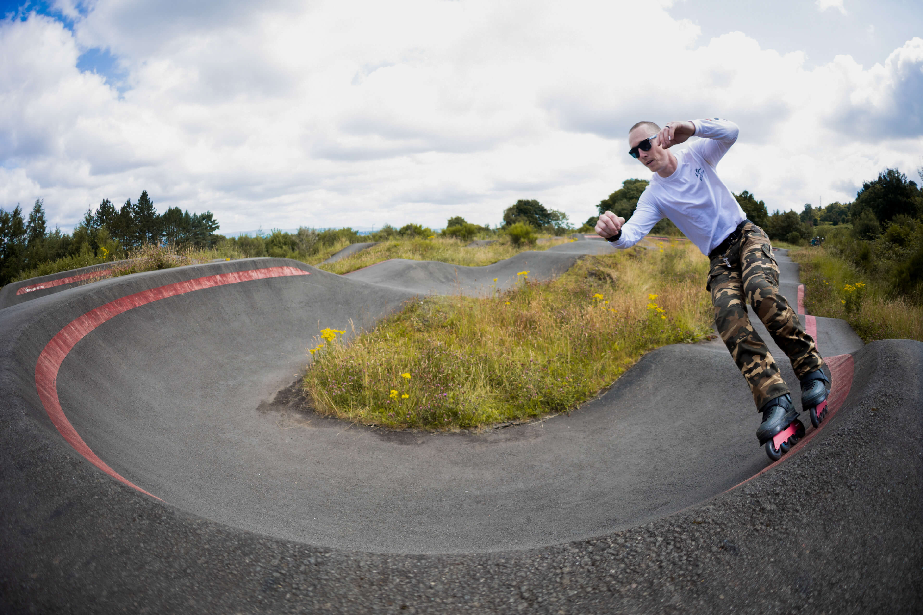 A man wearing a white long-sleeve shirt, camouflage pants, and inline skates rides around a curved asphalt pump track with red painted edges in an outdoor park setting.