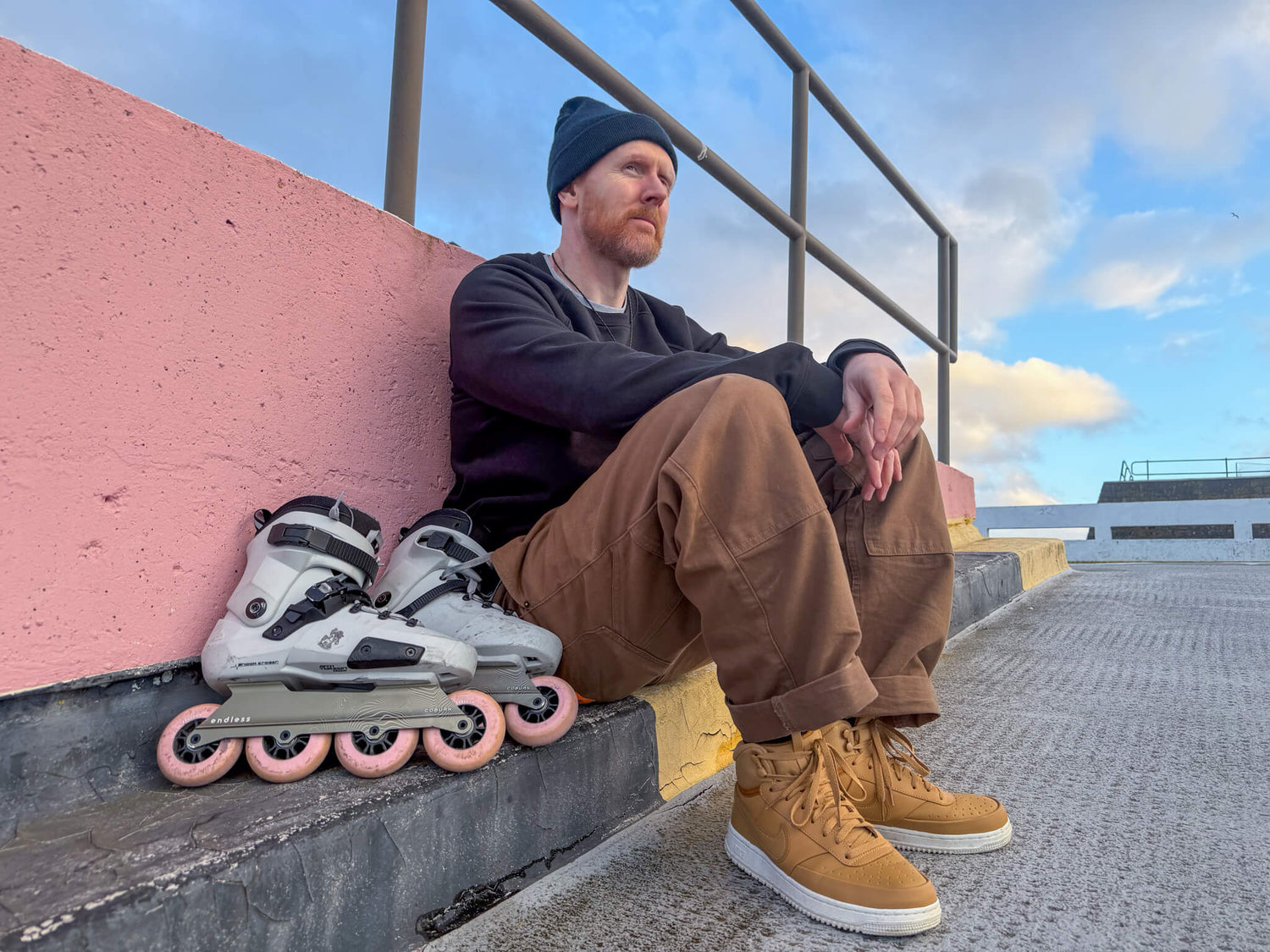 Si Coburn sitting on a ledge with Endless Coburn skate setup next to them, wearing a black beanie and brown pants.