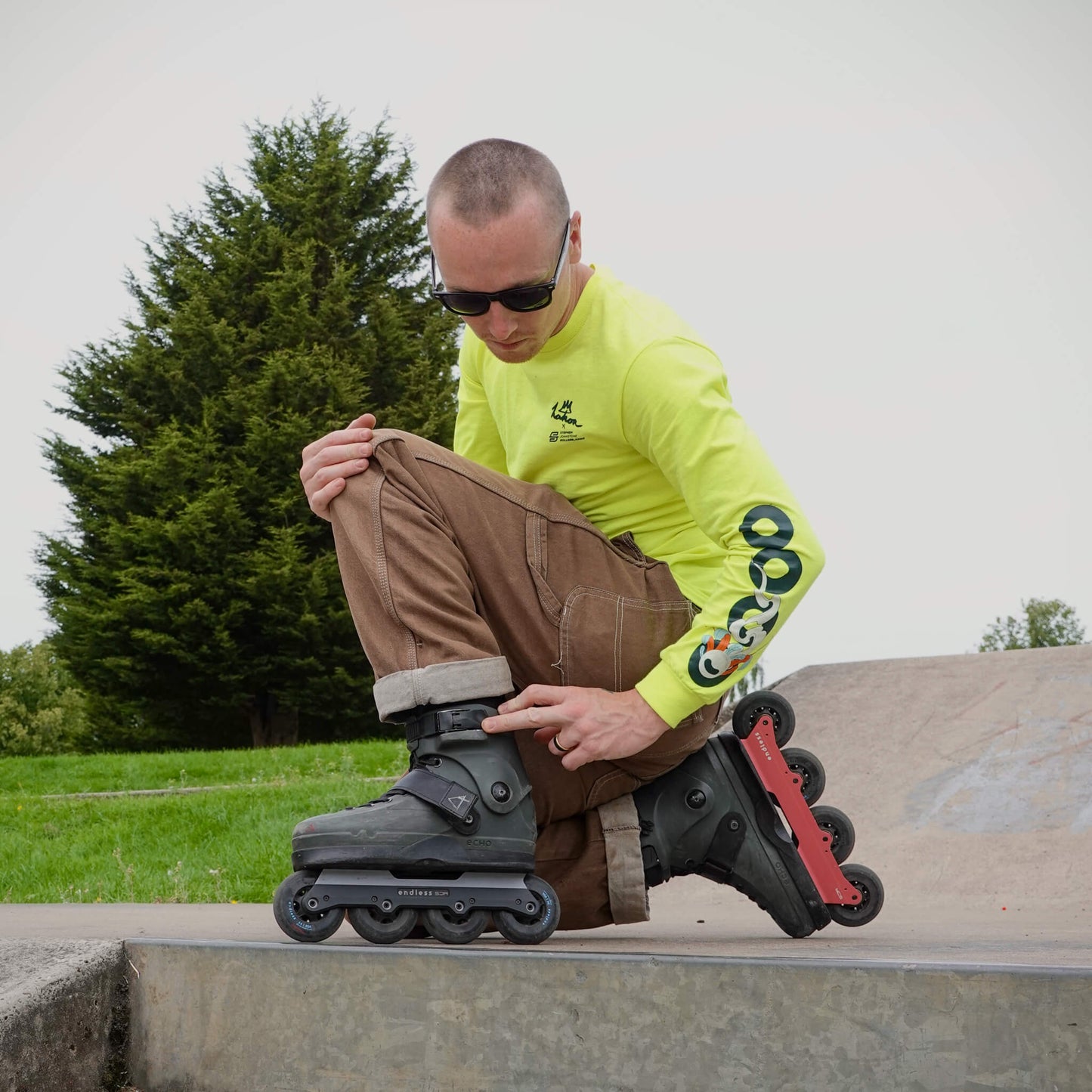 A skater wearing a neon yellow long-sleeve shirt kneeling to adjust his Endless Blading Co. SJR UFS light red inline frames.